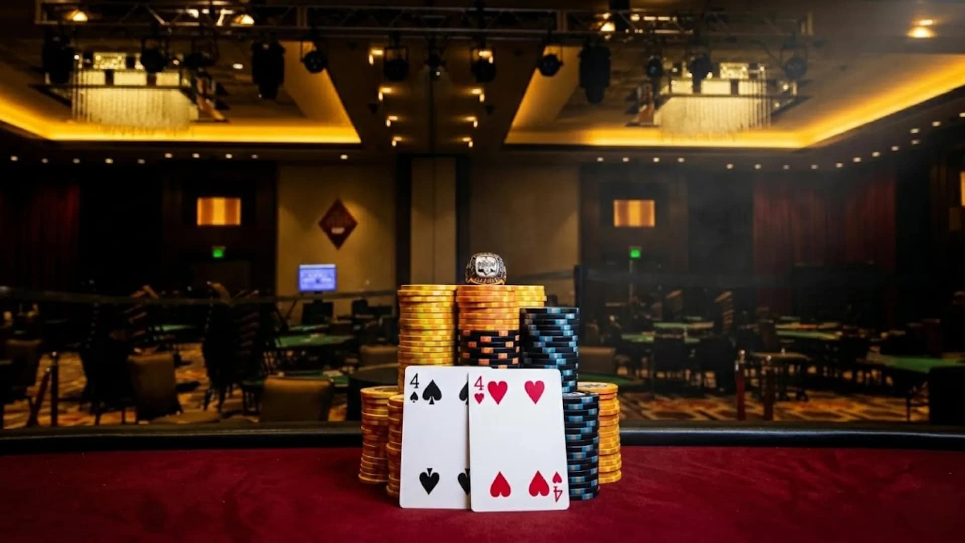 Close-up of a high-stakes poker winner's setup featuring massive stacks of yellow and blue chips, a four of spades and four of hearts, and a coveted detailed champion ring on top, in a luxurious casino setting.